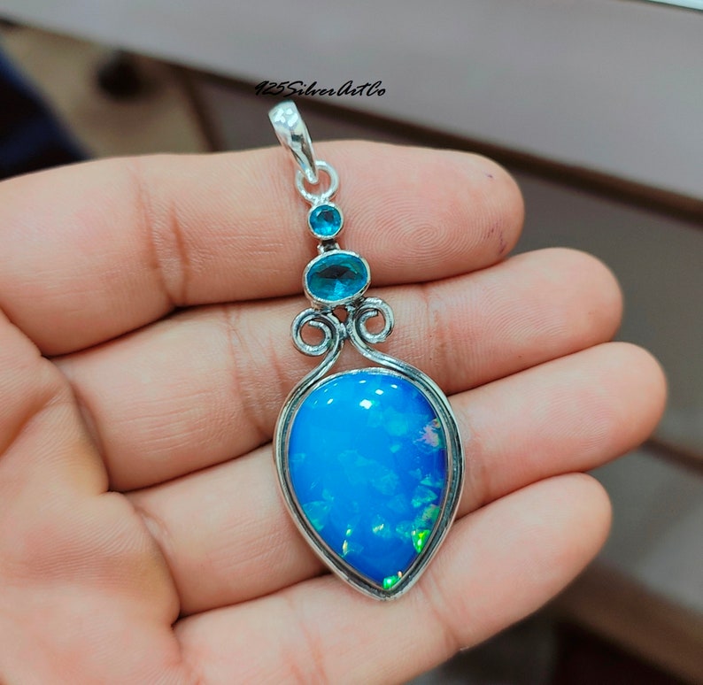 May include: A silver pendant with a blue opal gemstone and two smaller blue gemstones. The pendant has a delicate, ornate design.
