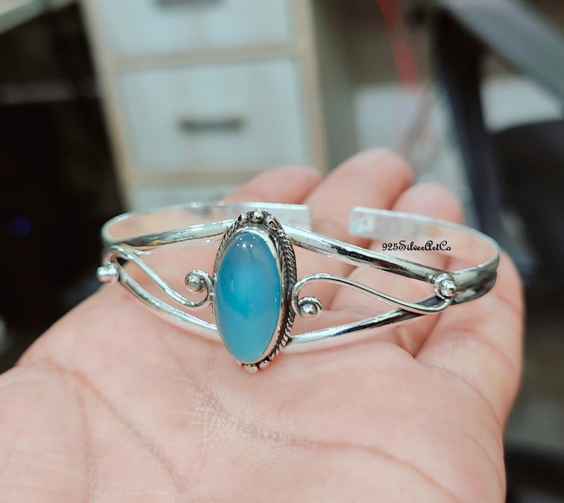 K&ouml;nnte beinhalten: Ein silbernes Armreif-Armband mit einem hellblauen ovalen Edelstein, der in der Mitte eingesetzt ist. Das Armband hat ein zartes, verziertes Design mit zwei geschwungenen B&auml;ndern und einer kleinen Perle auf jeder Seite des Edelsteins.