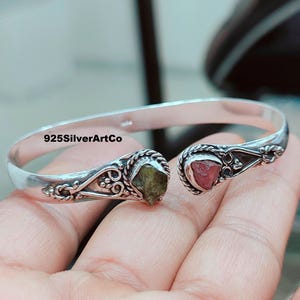 May include: Silver bangle bracelet with a green and pink gemstone accent. The bracelet is open and has a delicate design.