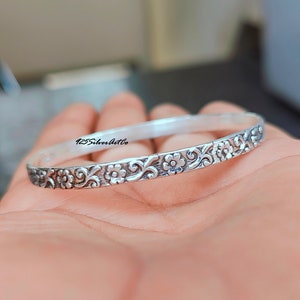 Op de afbeelding: Een zilveren armband met een bloemenpatroon. De armband is versierd met een herhalend ontwerp van bloemen en wervelende ranken. De tekst "425SilverArtCo" is zichtbaar op de armband.