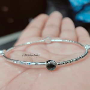 Op de afbeelding: Zilveren armband met een gehammerde afwerking en vier blauwe edelstenen. De armband is op een hand, en de tekst "925SilverArtCo" is zichtbaar op de armband.