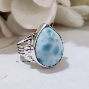 Puede incluir: Un anillo de plata con una piedra preciosa azul y blanca en forma de lágrima. El anillo tiene un diseño delicado con una banda que se envuelve alrededor del dedo.