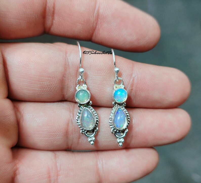 May include: A pair of silver dangle earrings. Each earring features a round, light-colored stone above a pear-shaped, iridescent stone. The earrings have a decorative, beaded border and are suspended from a simple hook. The text "125SilverArtCo" is visible.