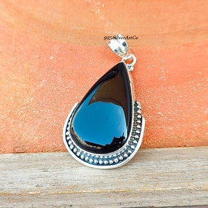 May include: A black gemstone pendant with a silver setting. The pendant is shaped like a teardrop and has a beaded border. The pendant is on a brown surface.
