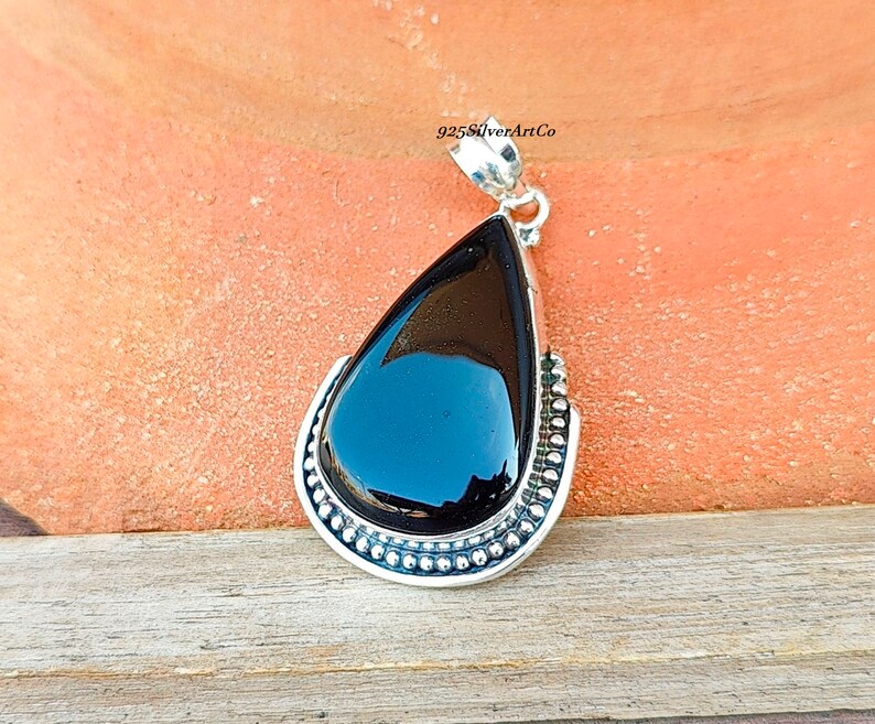 May include: A black gemstone pendant with a silver setting. The pendant is shaped like a teardrop and has a beaded border. The pendant is on a brown surface.