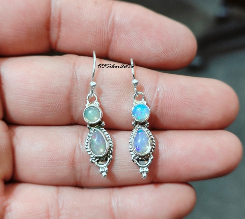 May include: A pair of silver drop earrings featuring two opalescent stones. The top stone is round, and the bottom stone is teardrop-shaped, with a decorative silver border. The earrings are suspended from a simple hook.