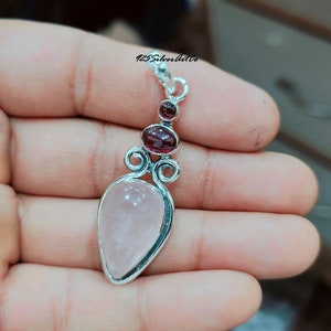 May include: A silver pendant with a pink rose quartz stone and two red garnet stones. The pendant has a delicate, ornate design.