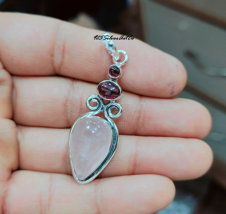 May include: A silver pendant with a pink rose quartz stone and two red garnet stones. The pendant has a delicate, ornate design.