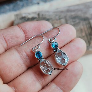 May include: Silver dangle earrings with a clear quartz teardrop and a small blue gemstone. The earrings have a hook closure.