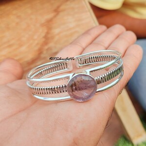 May include: A silver bangle bracelet with a large, round, light purple amethyst gemstone set in the center. The bracelet has a textured, braided design.