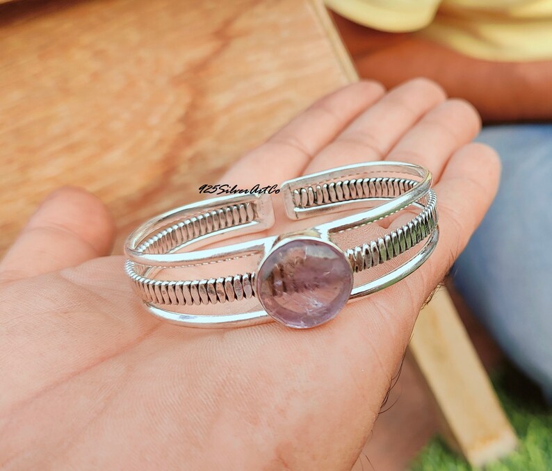 May include: A silver bangle bracelet with a large, round, light purple amethyst gemstone set in the center. The bracelet has a textured, braided design.