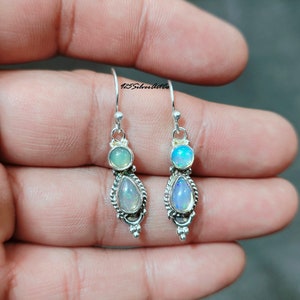 May include: A pair of silver dangle earrings. Each earring features a round, light-colored stone above a pear-shaped, iridescent stone. The earrings have a decorative, beaded border and are suspended from a simple hook. The text "125SilverArtCo" is visible.