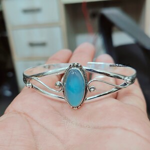 K&ouml;nnte beinhalten: Silbernes Armreif-Armband mit einem hellblauen ovalen Edelstein. Das Armband hat ein zartes, geschwungenes Design.