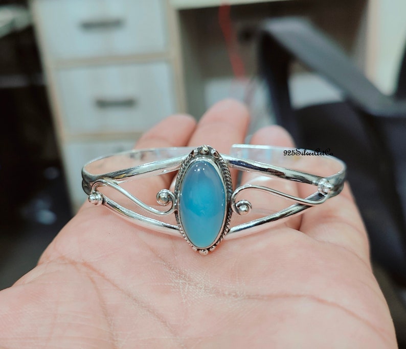 K&ouml;nnte beinhalten: Silbernes Armreif-Armband mit einem hellblauen ovalen Edelstein. Das Armband hat ein zartes, geschwungenes Design.
