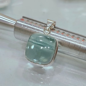 Könnte beinhalten: Ein silberner Anhänger mit einem hellblauen Aquamarin-Edelstein. Der Anhänger ist quadratisch und hat eine glatte, polierte Oberfläche. Der Edelstein ist in einer silbernen Fassung gefasst.