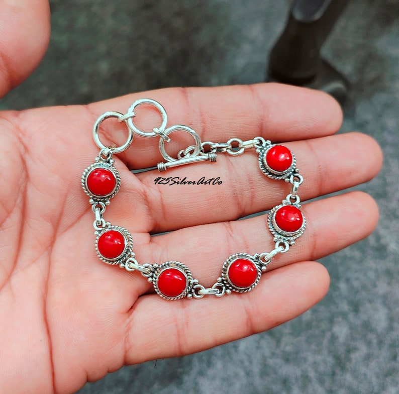 May include: A silver bracelet with a toggle clasp and six red gemstone beads. The bracelet is on a hand.