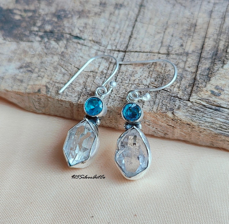 May include: Silver dangle earrings with a clear quartz crystal and a blue gemstone. The earrings have a simple design and are perfect for everyday wear.