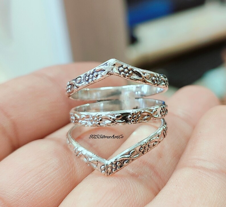 Op de afbeelding: Een zilveren ring met een bloemenmotief. De ring is gemaakt van drie banden die met elkaar zijn verweven. De banden zijn V-vormig. De ring zit aan een vinger.