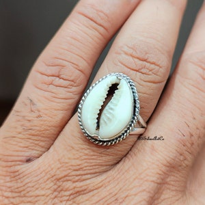 Puede incluir: Un anillo de plata con una concha de cauri blanca engastada en un diseño de cuerda. El anillo está en un dedo.