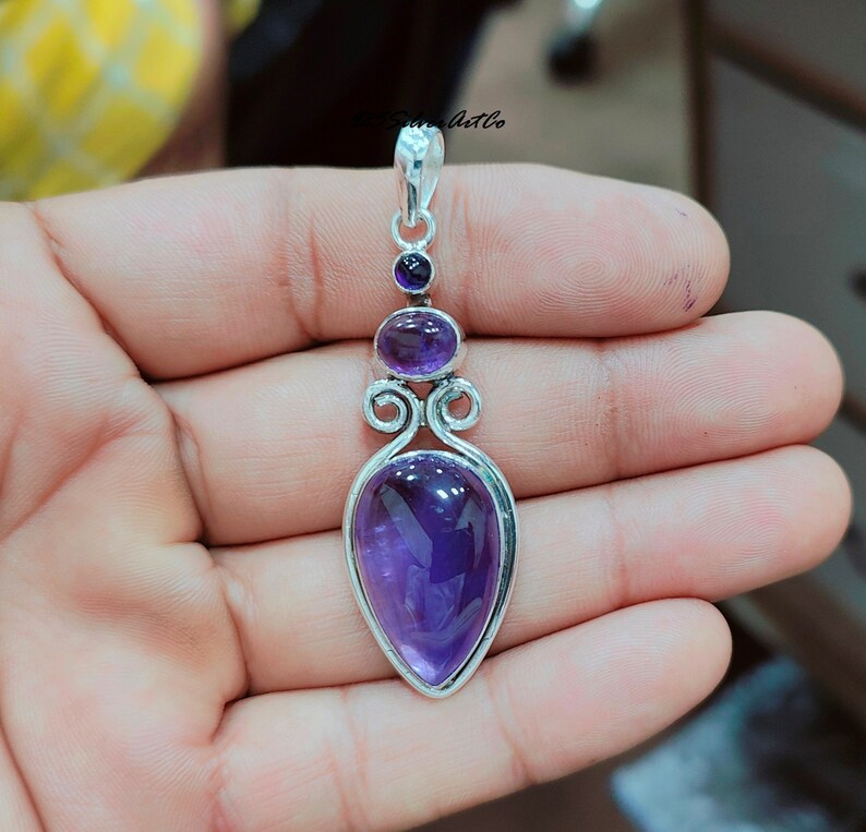 May include: A silver pendant with a teardrop-shaped amethyst gemstone. The pendant has a delicate design with two smaller amethyst gemstones above the main stone.