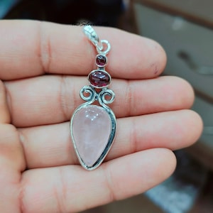 May include: A silver pendant with a pink rose quartz stone and two red garnet stones. The pendant is in the shape of a teardrop.