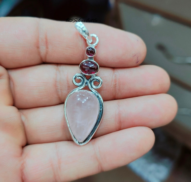 May include: A silver pendant with a pink rose quartz stone and two red garnet stones. The pendant is in the shape of a teardrop.