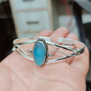 K&ouml;nnte beinhalten: Ein silbernes Armreif-Armband mit einem hellblauen ovalen Edelstein, der in der Mitte eingesetzt ist. Das Armband hat ein zartes, verziertes Design mit zwei geschwungenen B&auml;ndern und einer kleinen Perle auf jeder Seite des Edelsteins.
