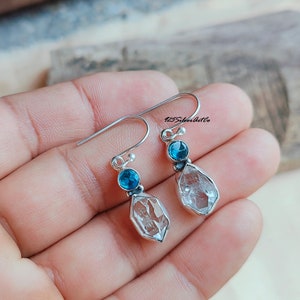 May include: Silver dangle earrings with a clear quartz crystal and a blue topaz gemstone. 125SilverArtCo is printed on the image.