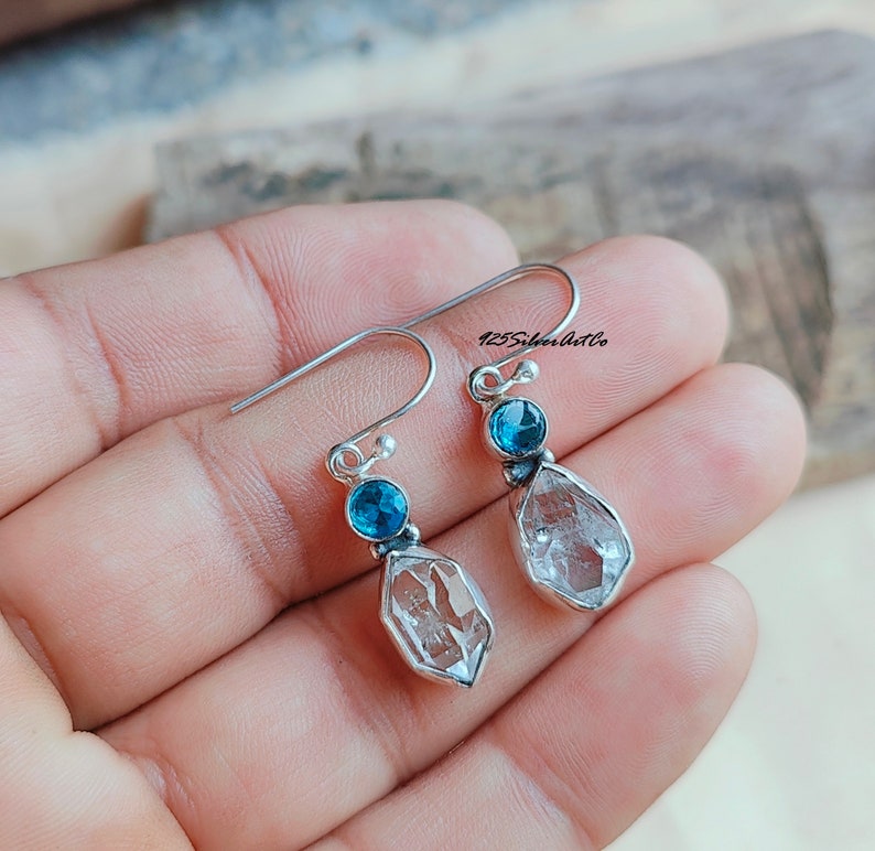 May include: Silver dangle earrings with a clear quartz crystal and a blue topaz gemstone. 125SilverArtCo is printed on the image.