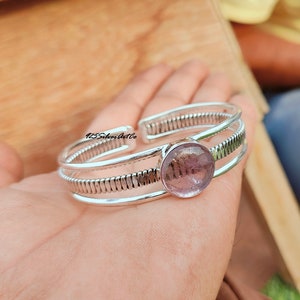 May include: Silver bangle bracelet with a round, light purple gemstone. The bracelet has a textured, ribbed design.