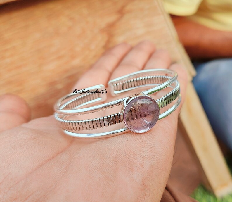 May include: Silver bangle bracelet with a round, light purple gemstone. The bracelet has a textured, ribbed design.