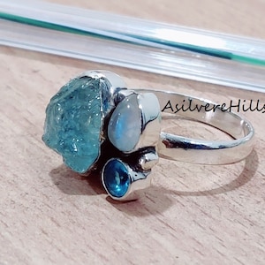 Pode incluir: Um anel de prata com três pedras preciosas: uma grande água-marinha, uma pedra da lua menor e uma topázio azul. O anel é engastado em uma banda de prata e tem o texto "AsilverHills" gravado na lateral.