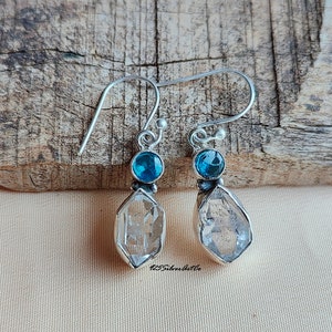 May include: A pair of silver earrings with a clear quartz crystal and a blue gemstone. The earrings have a simple design and are perfect for everyday wear.