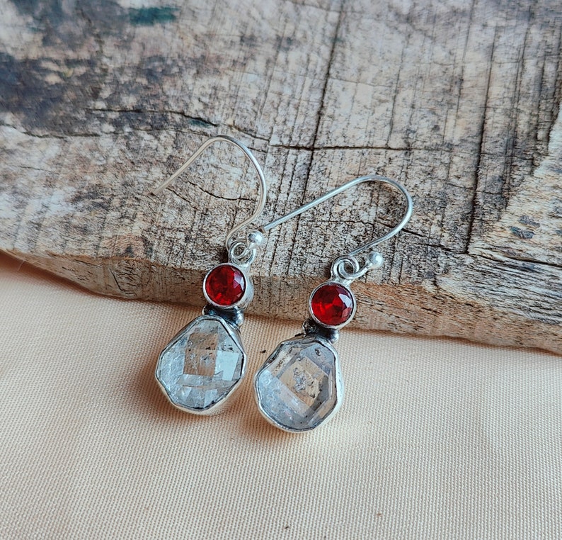 May include: A pair of silver earrings with clear faceted stones and red gemstone accents.