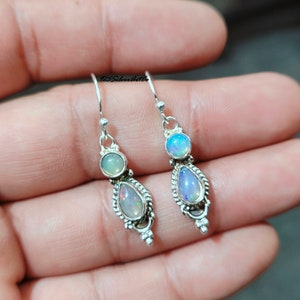 May include: A pair of silver earrings with opal gemstones. Each earring features a teardrop-shaped opal, a round opal, and a decorative silver setting. The earrings are displayed against a neutral background.