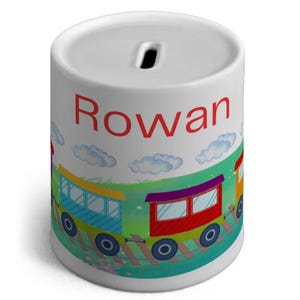 Puede incluir: Huchita de cerámica blanca con un diseño de tren. El tren está formado por tres vagones, uno rojo, uno azul y uno amarillo. El tren está en una vía con hierba verde y nubes blancas. El nombre "Rowan" está escrito en letras rojas en la parte delantera de la huchita.