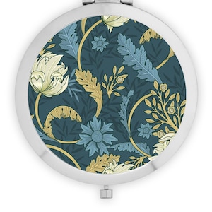 May include: A round silver compact mirror with a floral design. The design features blue, yellow, and white flowers on a teal background.