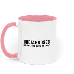May include: White ceramic coffee mug with a pink rim and handle. The mug has black text that reads "Undiagnosed but something deffo isn't right."
