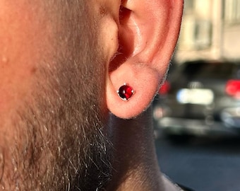 Puede incluir: Primer plano de una oreja con un pequeño pendiente de botón de piedra preciosa roja y redonda. El pendiente está engastado en metal plateado o blanco. La persona lleva una camiseta negra y gafas de sol.