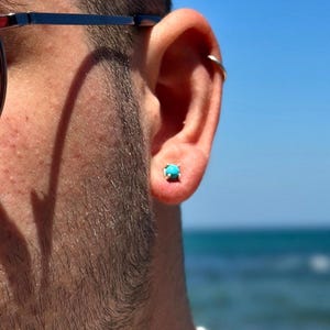 Puede incluir: Primer plano de una persona con gafas de sol redondas negras y un collar de perlas. Un pequeño pendiente turquesa es visible en la oreja. El fondo muestra un cielo azul claro y el mar.