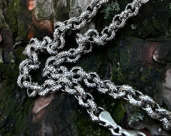 Puede incluir: Un collar de cadena de plata con un diseño detallado e intrincado. La cadena está sobre un tronco de árbol cubierto de musgo.