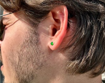 May include: A close-up of a person's ear with a small, square-cut, bright green gemstone stud earring. The earring is set in a gold-colored metal. The person is wearing sunglasses and has brown hair and a beard.