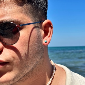 Puede incluir: Un hombre con una camisa blanca, un collar de perlas y gafas de sol. Lleva un pendiente de piedra turquesa en la oreja derecha.