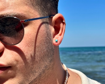 Puede incluir: Un hombre con una camisa blanca, un collar de perlas y gafas de sol. Lleva un pendiente de piedra turquesa en la oreja derecha.