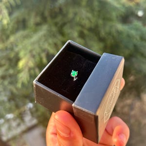Puede incluir: Una peque&ntilde;a caja de joyer&iacute;a negra abierta, que revela un solo pendiente de esmeralda. El pendiente tiene una piedra preciosa verde brillante y un engaste plateado. La caja se sostiene en una mano, con un fondo verde borroso.