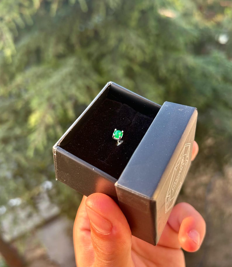 Puede incluir: Una peque&ntilde;a caja de joyer&iacute;a negra abierta, que revela un solo pendiente de esmeralda. El pendiente tiene una piedra preciosa verde brillante y un engaste plateado. La caja se sostiene en una mano, con un fondo verde borroso.