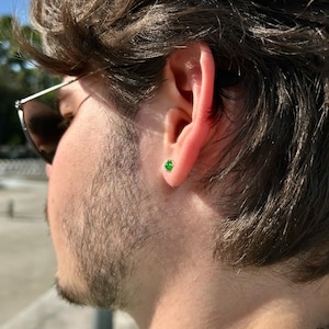 Puede incluir: Primer plano de una oreja con un peque&ntilde;o pendiente de bot&oacute;n redondo de color verde esmeralda. El pendiente est&aacute; engastado en oro o plata. La persona lleva gafas de sol y tiene el pelo oscuro y barba. Una joya cl&aacute;sica.