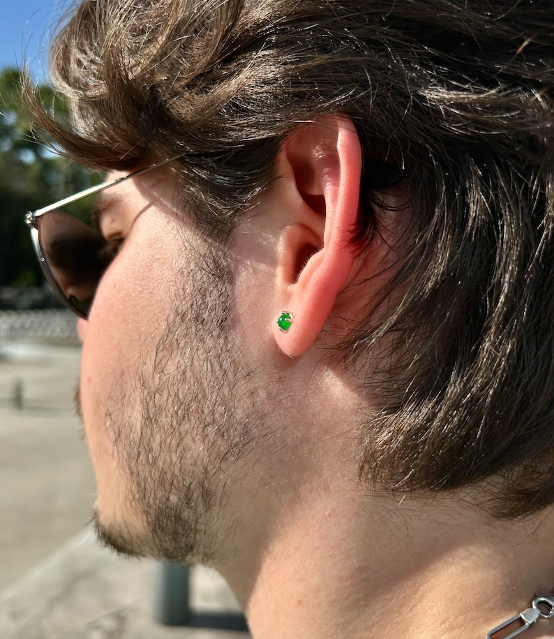 Puede incluir: Primer plano de una oreja con un peque&ntilde;o pendiente de bot&oacute;n redondo de color verde esmeralda. El pendiente est&aacute; engastado en oro o plata. La persona lleva gafas de sol y tiene el pelo oscuro y barba. Una joya cl&aacute;sica.