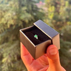 Puede incluir: Un peque&ntilde;o anillo con una piedra preciosa de color esmeralda en una caja de joyer&iacute;a negra. El anillo est&aacute; engastado en una banda plateada y se muestra sobre un interior de terciopelo negro. La caja est&aacute; abierta.