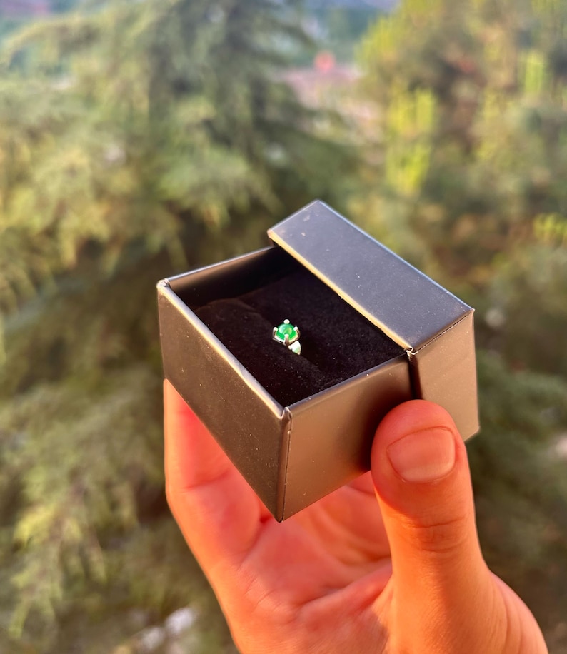 Puede incluir: Un peque&ntilde;o anillo con una piedra preciosa de color esmeralda en una caja de joyer&iacute;a negra. El anillo est&aacute; engastado en una banda plateada y se muestra sobre un interior de terciopelo negro. La caja est&aacute; abierta.